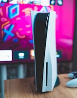 A gaming console is placed on a wooden table with a game controller nearby. In the background, a television screen displays colorful graphics, including geometric shapes and vibrant colors.