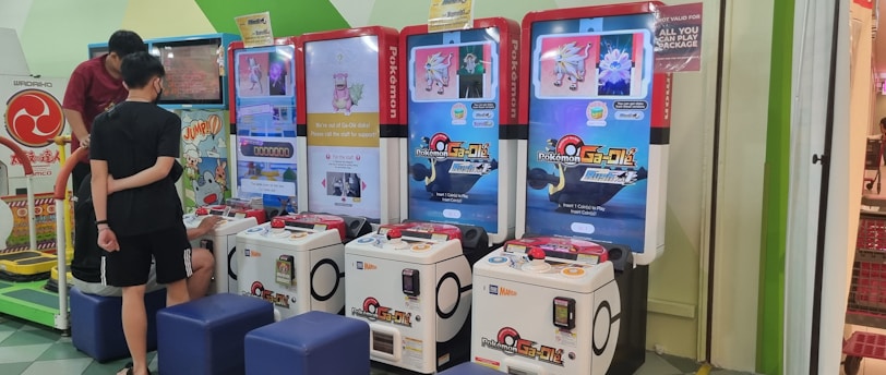 Several arcade gaming machines with multiple screens displaying Pokémon-themed content are positioned in a colorful arcade setting. There are people interacting with the machines; some are seated, and others are standing. The area features different gaming options, including a jumping game with bright, inviting graphics.